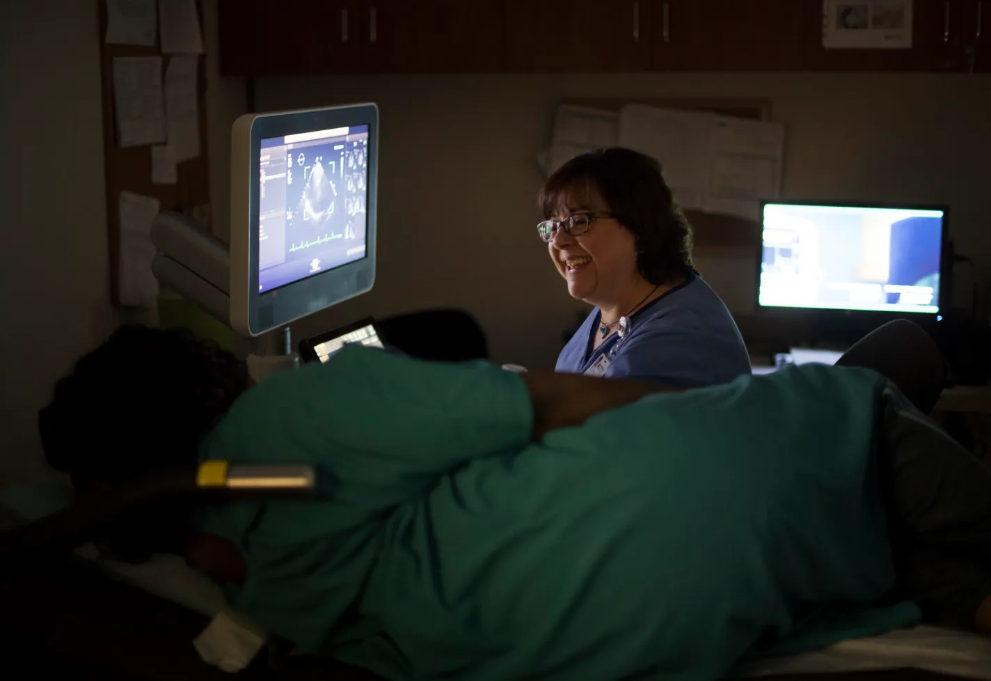 A Novant Health imagining tech is sitting at an ultrasound machine talking and smiling with a patient, who is laying on a table. 
