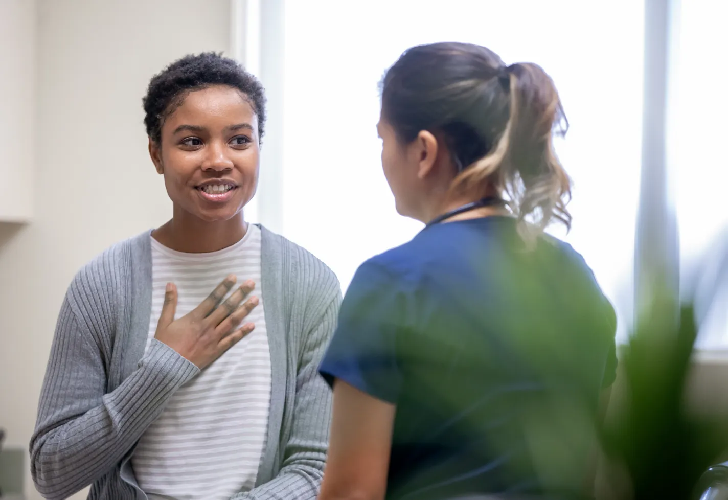 A young woman is sitting in an exam room talking with a nurse. As she talks, she has placed her hand over her chest. 