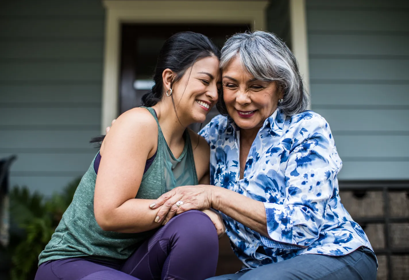 A mother, and her adult daughter, are sitting on their front porch smiling and hugging one another. 