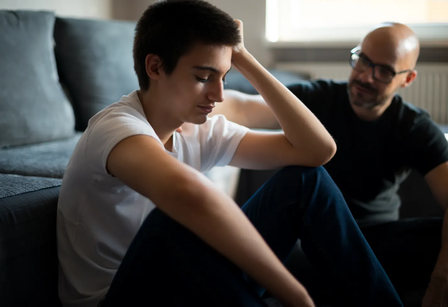 A teenage boy is resting his head on his hand as he sits on the floor and talks with his father. 