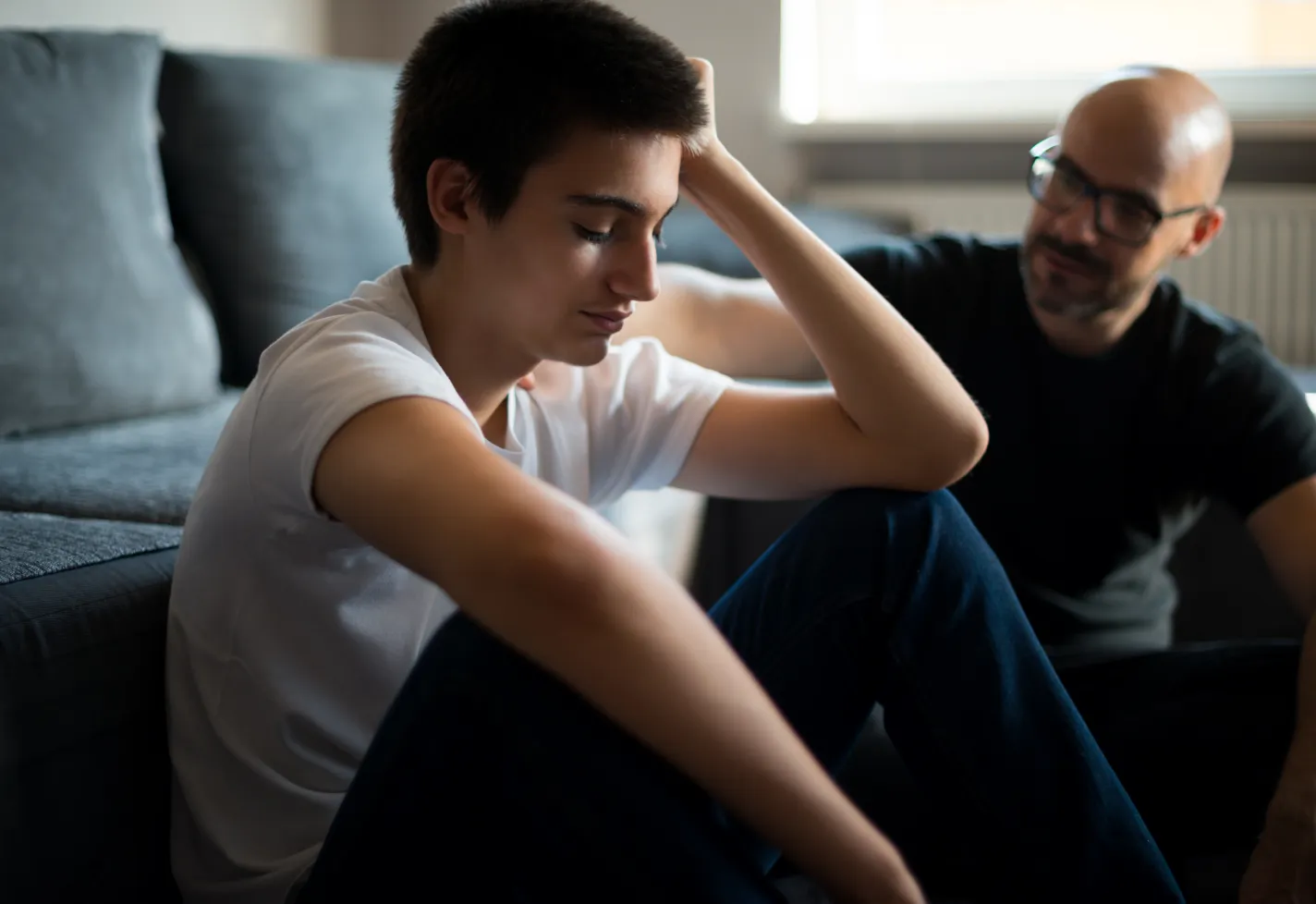 A teenage boy is resting his head on his hand as he sits on the floor and talks with his father. A teenage boy is resting his head on his hand as he sits on the floor and talks with his father.