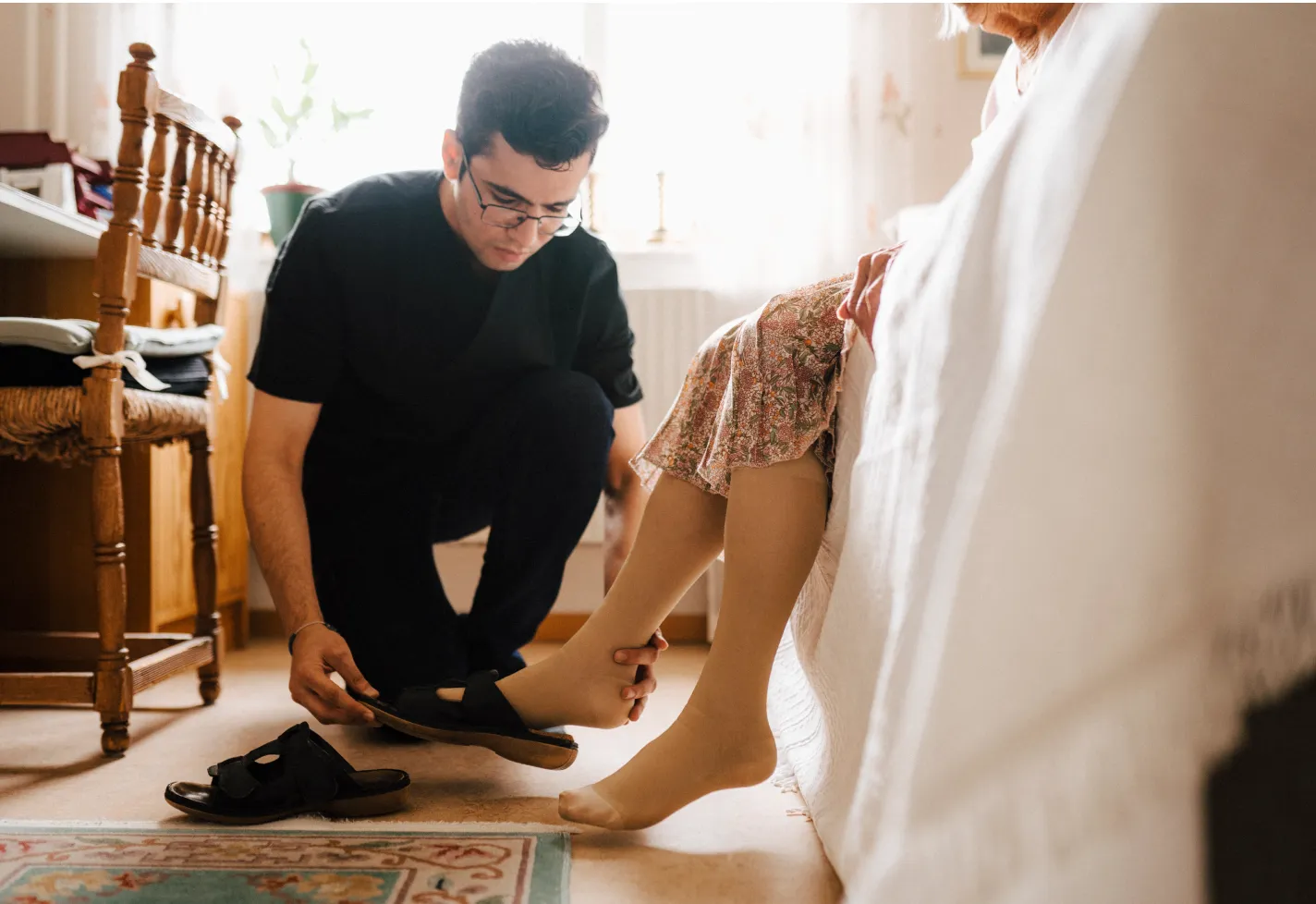 A caregiver is kneeling down to assist a senior woman by putting her shoes on. 