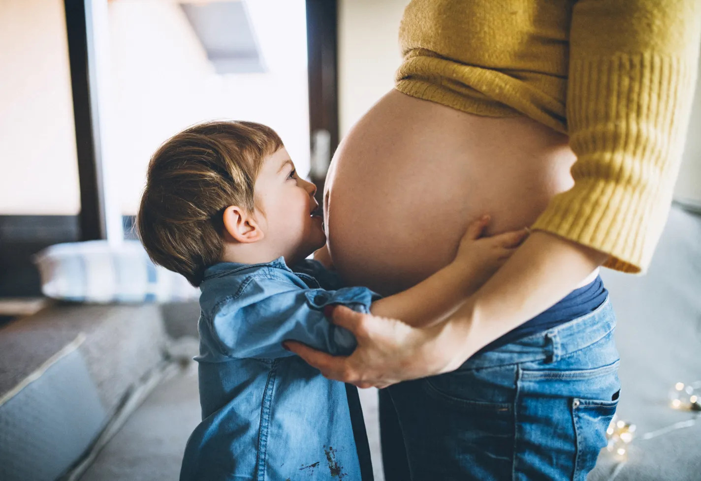 A pregnant woman is standing with her young child. The child is hugging and kissing his mother's baby bump. 