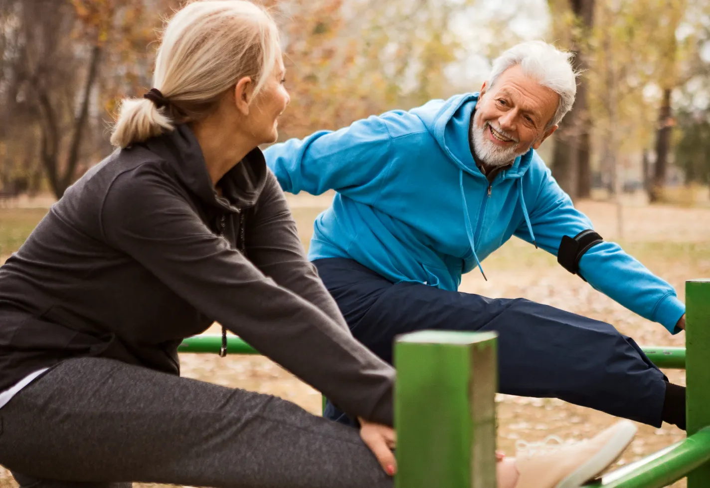 A senior couple is out at the park, in the fall. They both have one of their legs up on rails as they reach for their feet to stretch. 