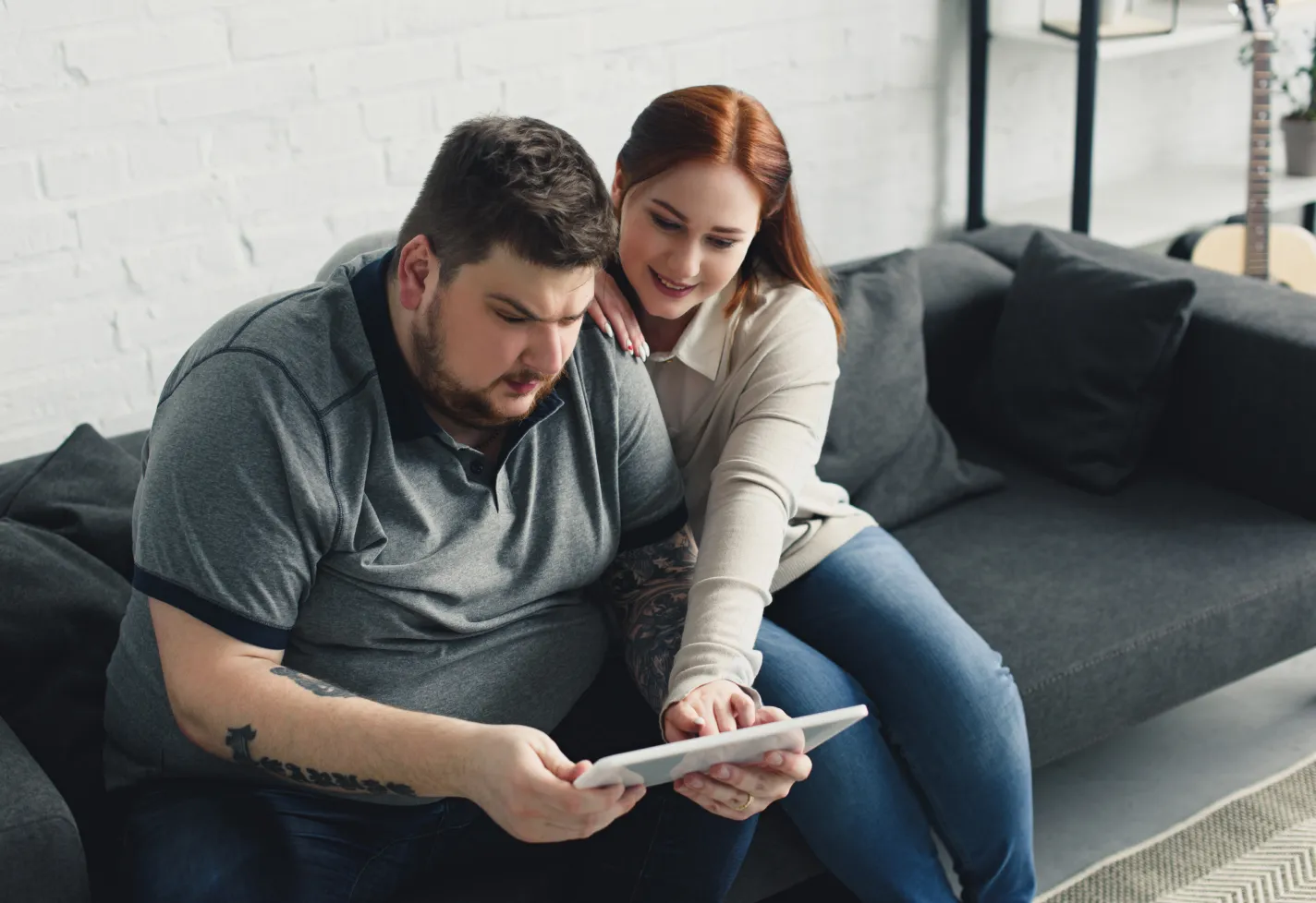 A man is sitting on the couch with his partner. He is holding a tablet and reviewing information, as his partner leans over to point at something on the screen. 