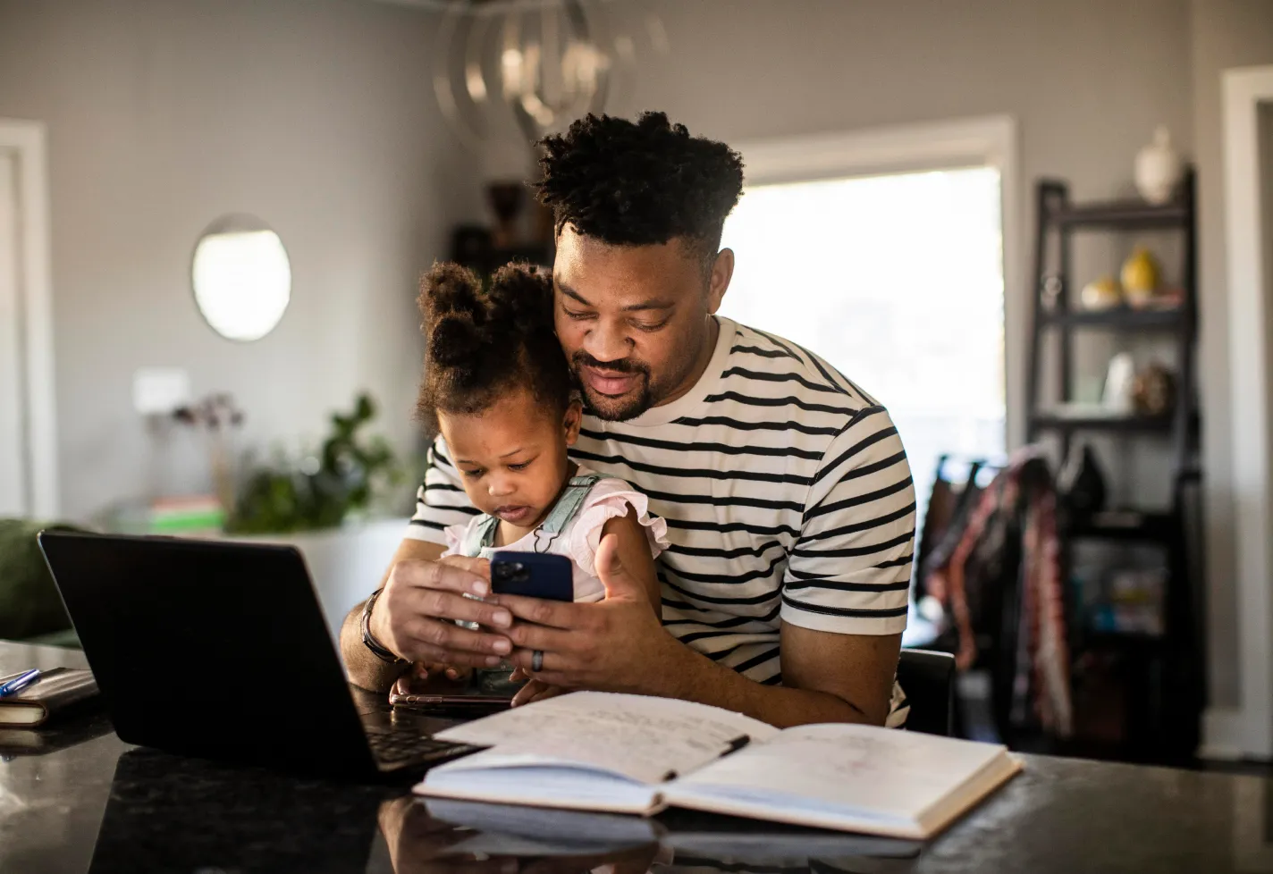 Father is sitting with his daughter at the counter. They are sitting in front of a laptop as he is looking for information on his phone. Father is sitting with his daughter at the counter. They are sitting in front of a laptop as he is looking for information on his phone.
