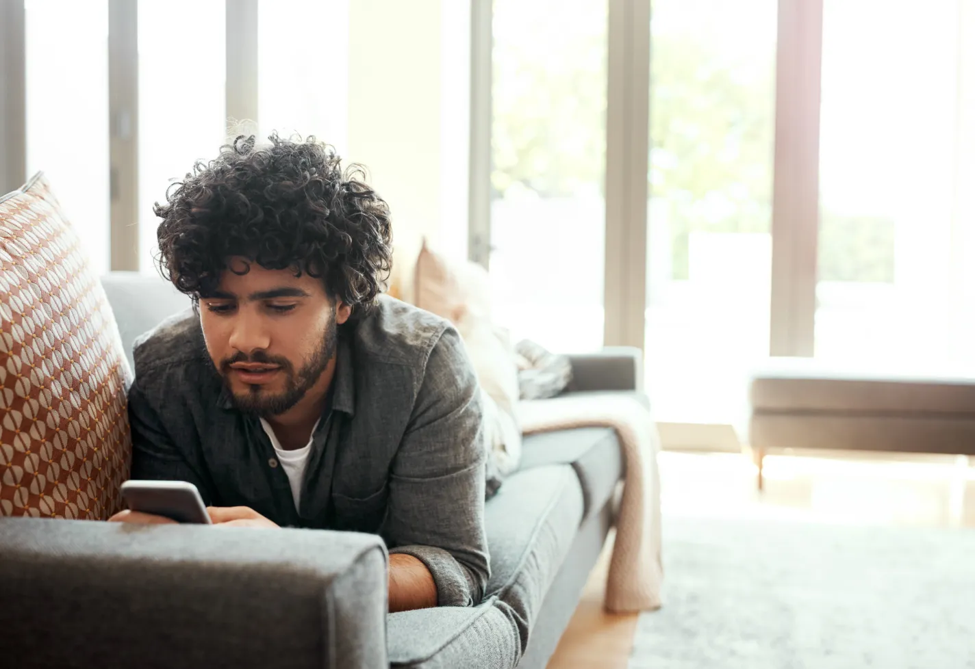 A young man is looking at information on his smartphone as he is lying down on a couch. 