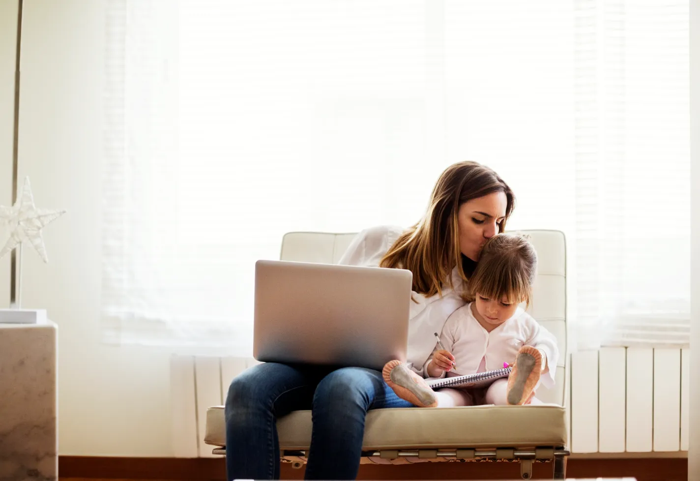 A mother is sitting next to her toddler with a laptop in her lap. The young girl is coloring on a pad of paper as her mother leans over to kiss her. 