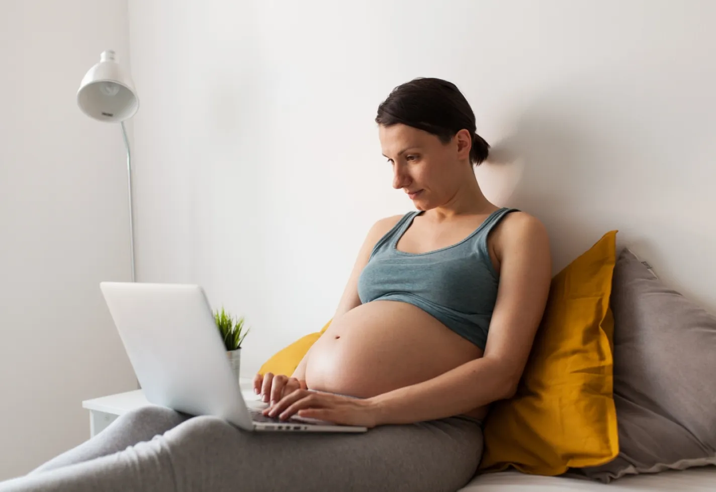 A pregnant woman is sitting in bed as she reviews information on a laptop. 