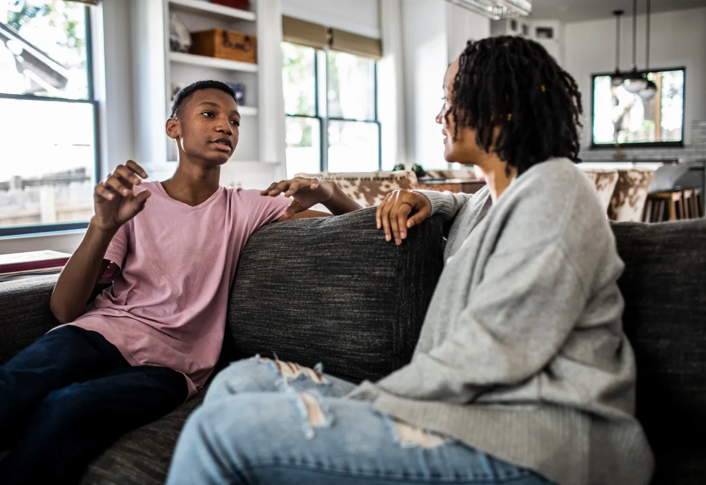 A son sits on his couch with his mom and uses his hand while talking with her A son sits on his couch with his mom and uses his hand while talking with her