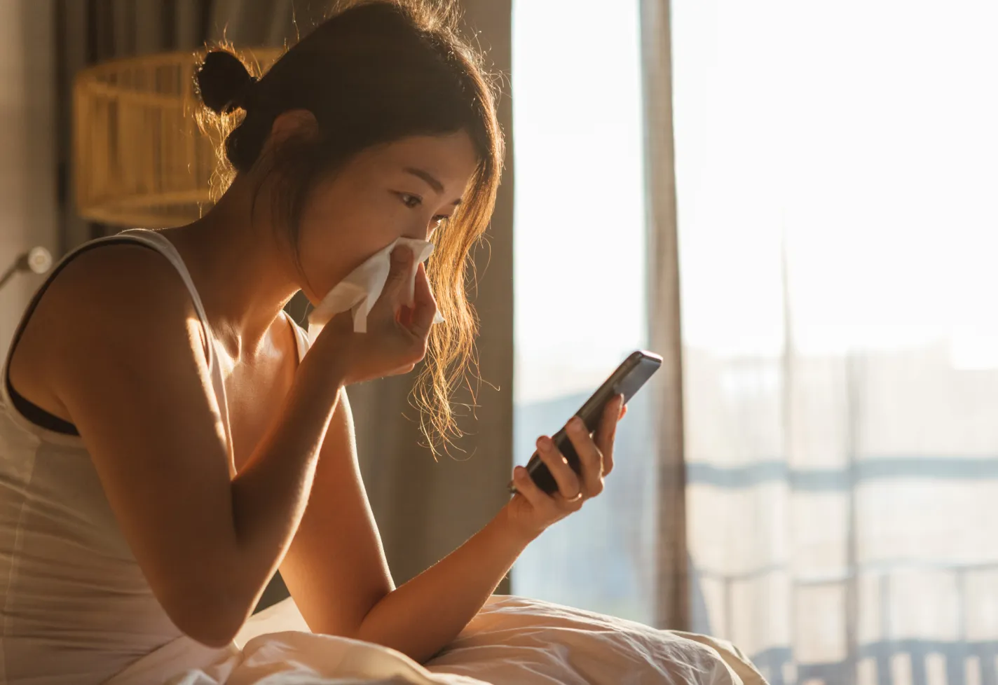 A young woman is sitting in her bed with a tissue at her nose. The sun is rising and she is reviewing information on her smart phone. 