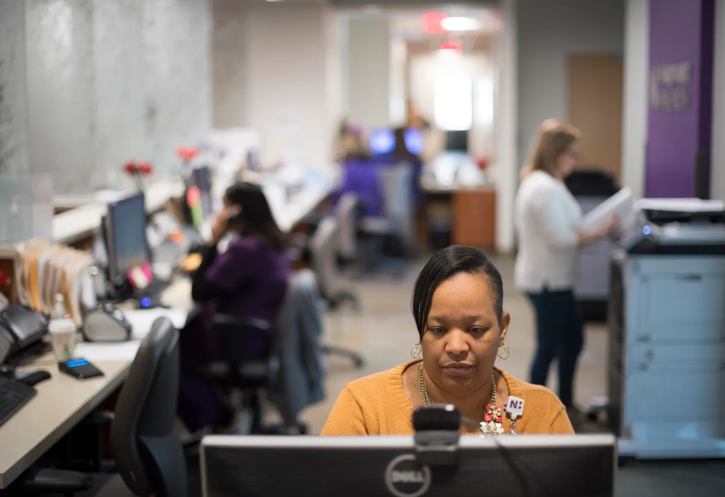 A Novant Health team member is sitting at her workstation on the computer while others are printing copies and on phone calls. 