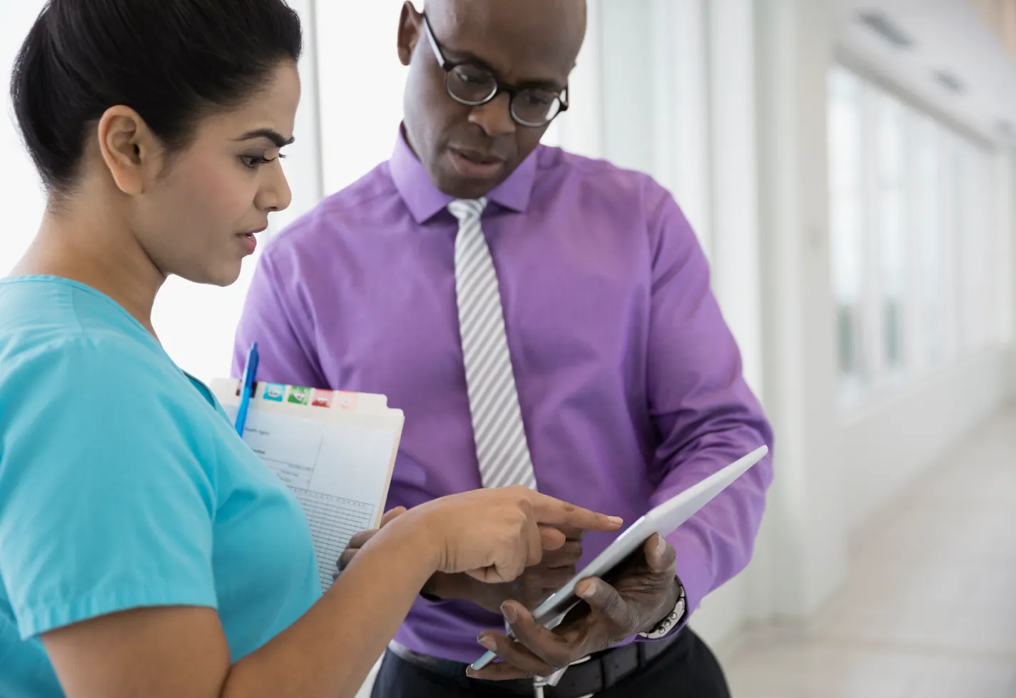 A healthcare provider is in a hallway reviewing patient information on a smart table as he talks with nurse. 