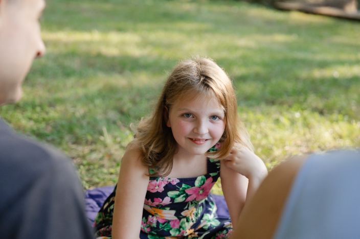 A girl with blonde hair wearing a floral dress, smiling while sitting on grass.