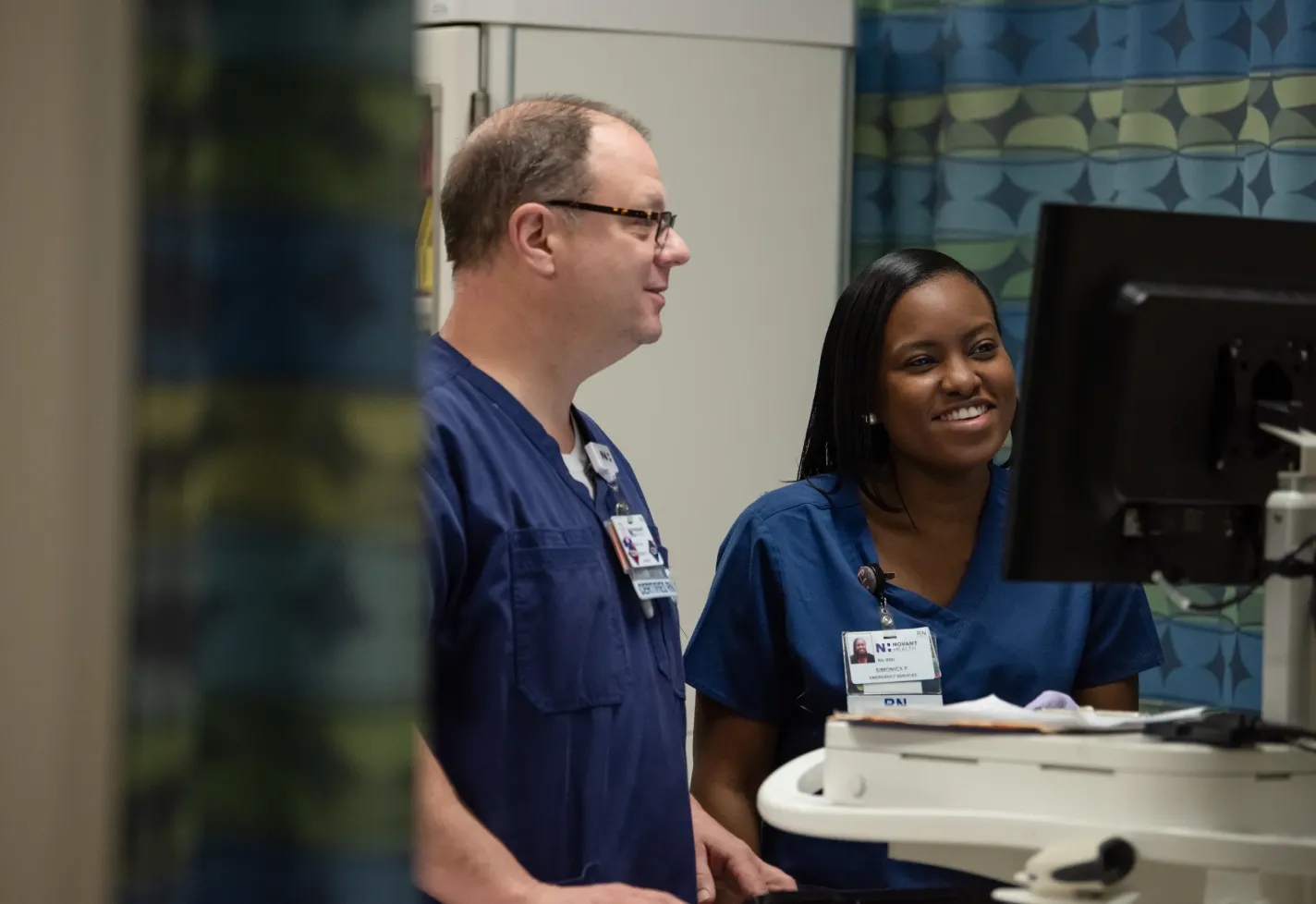 Two Novant Health nurses are standing together at a computer. One nurse is typing on the computer while the other is watching. 