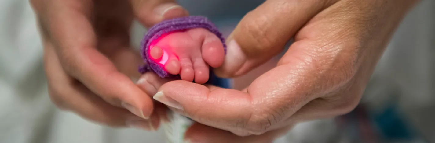 A health care provider has a newborns foot in their hand. The baby's foot has a red light senor on it that is wrapped in purple tape.