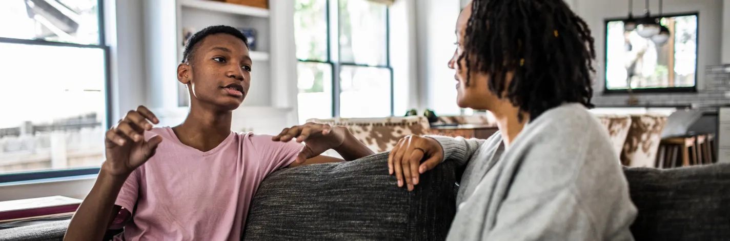 Young teenage boy at home, sitting on the couch, as he talks with his mother. 