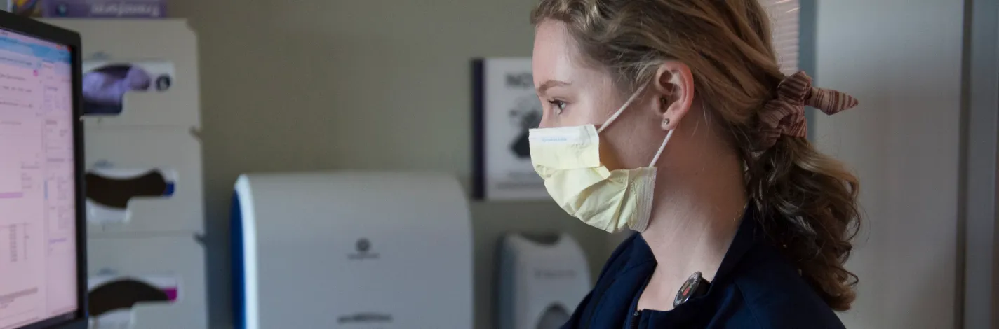 A masked nurse in patients room reviewing and updating the patients chart. 