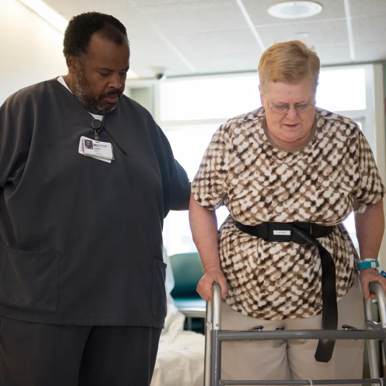 A male nurse is assistant a patient as she walks with a walker