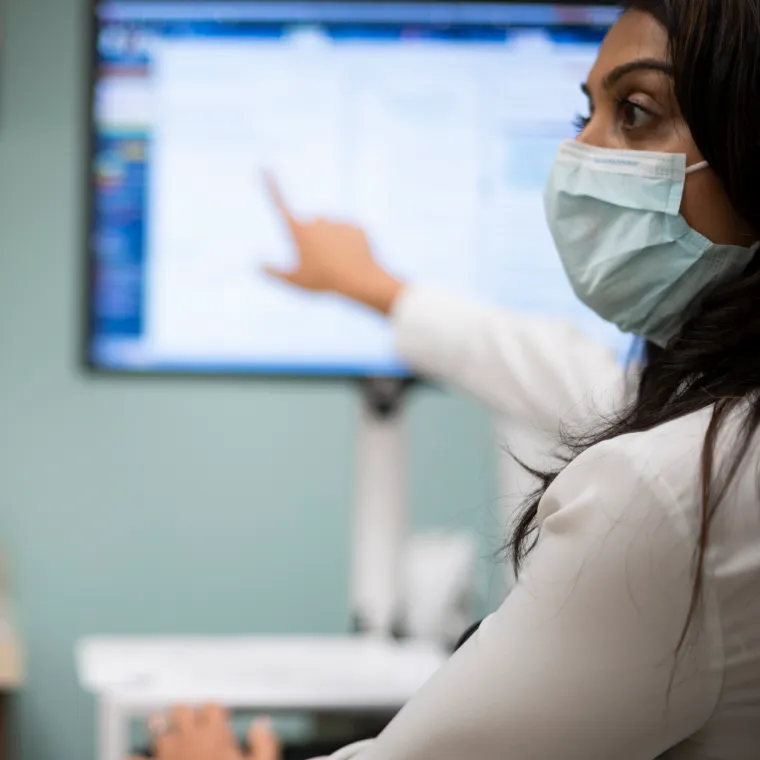 A healthcare provider is wearing a mask as she sits in the exam room and points at a computer screen. 
