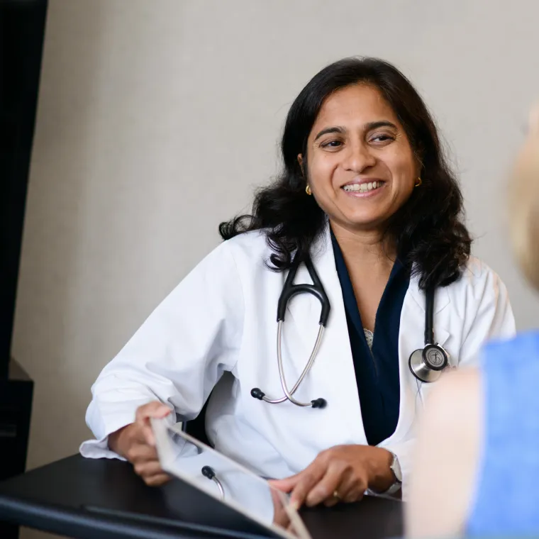 A healthcare provide is wearing a lab coat with stethoscope around their neck.  They are sitting in an exam room smiling at patient.