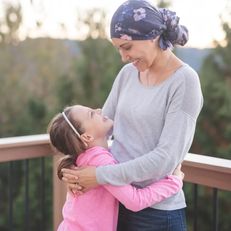 A young girl is looking up at her mother, who is wearing a headscarf due to cancer treatment. and smiling. Her mother is looking down at her and smiling back. A young girl is looking up at her mother, who is wearing a headscarf due to cancer treatment. and smiling. Her mother is looking down at her and smiling back.