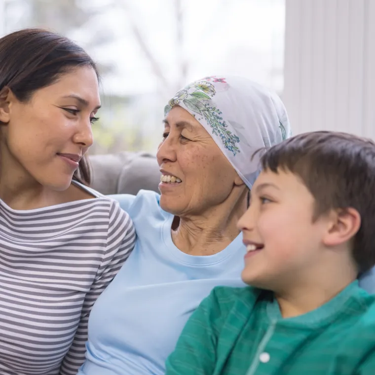 A grandmother is wearing a headscarf as she hugs and smiles at her adult daughter and grandchild. 