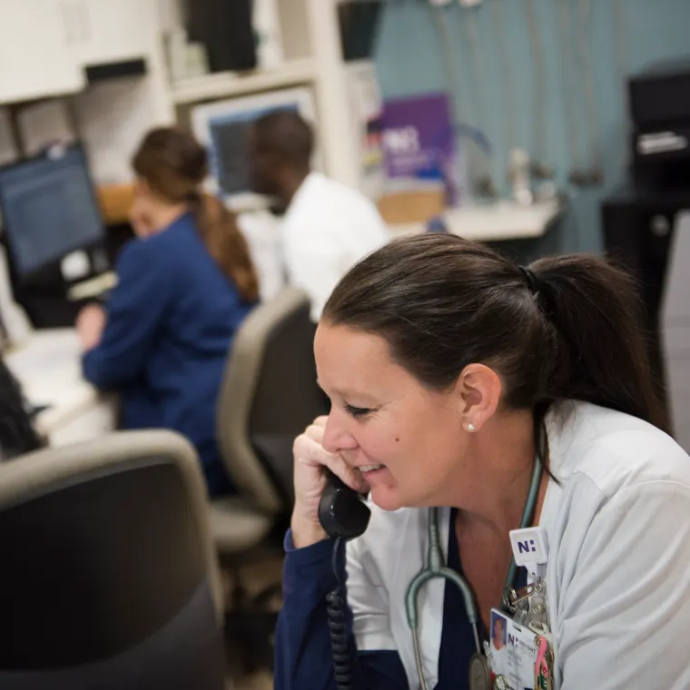 A Novant Health nurse is sitting at a nurse's station, in front of a computer, talking on the phone.