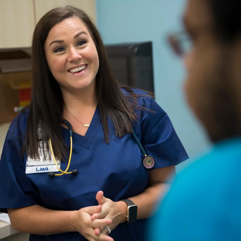 A Novant Health Certified Medical Assistant (CMA) is in an exam room talking and smiling with a male patient. 