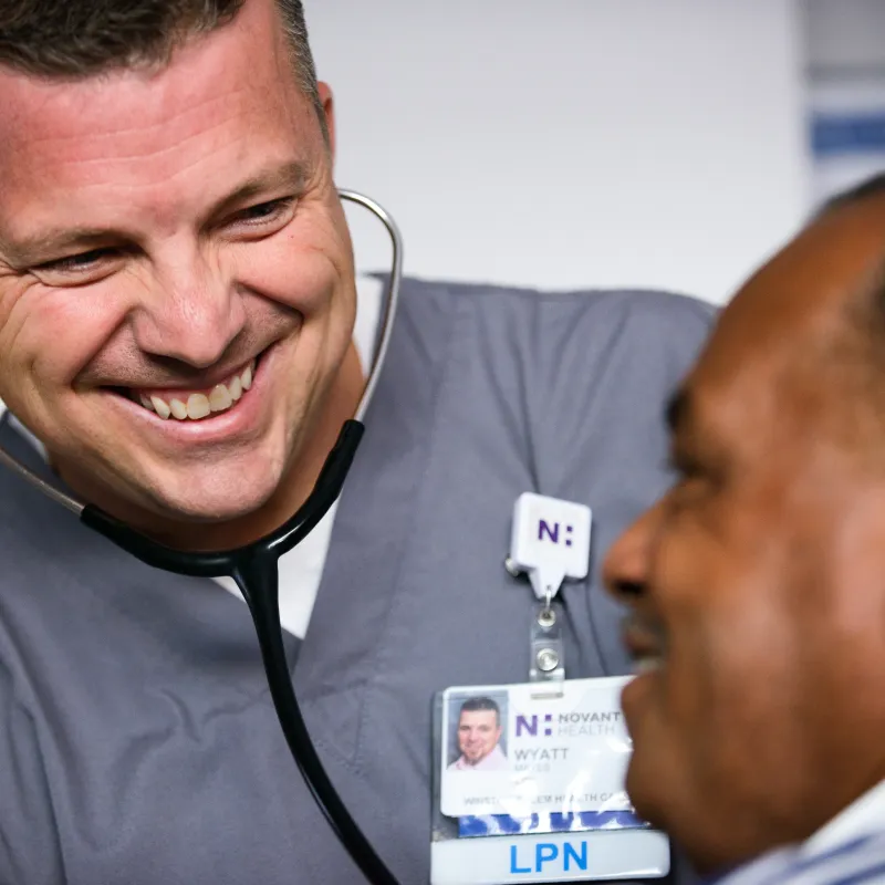 Novant Health nurse is in a clinic exam room with a patient. The nurse is wearing a stethoscope prepared to listen to the patient's heart but smiles as the patient is talking. 