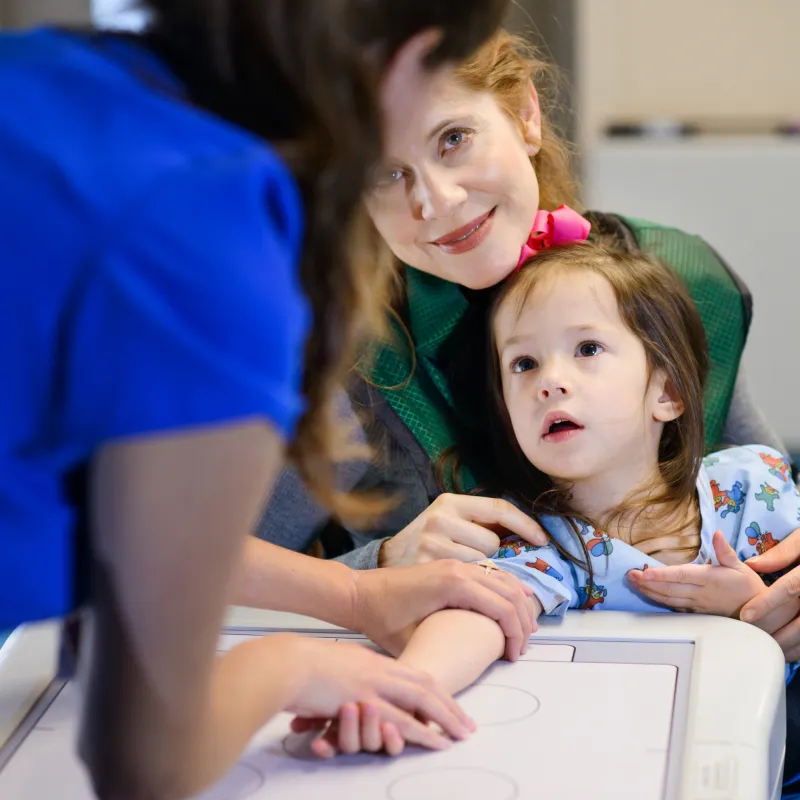 A young girl is sitting on her moms lap while a Novant Health team member positions her arm for an x-ray. 