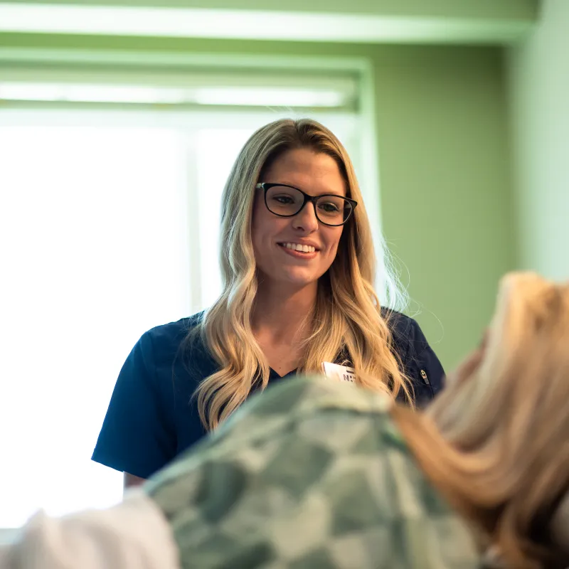 Novant Health nurse is standing in a patients recovery room. The nurse is smiling and talking with the patient as they lie in bed.