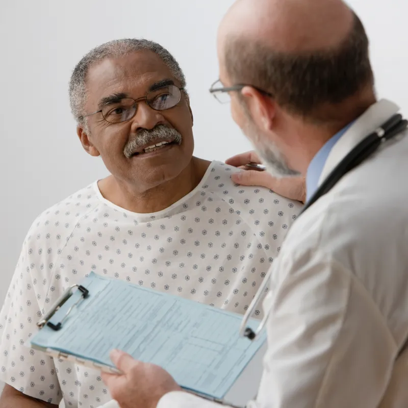 A senior man is talking with his doctor as he sits on the exam table in a gown. The doctor has placed his hand on the patients shoulder.