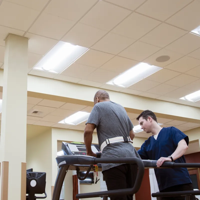 A man wearing a fitness belt walks on a treadmill while a physical therapist watches and supports him