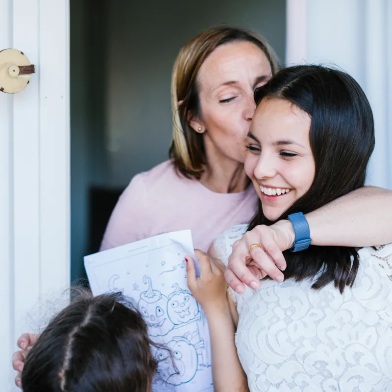 A teenager is smiling as her mom hugs and kisses her. 