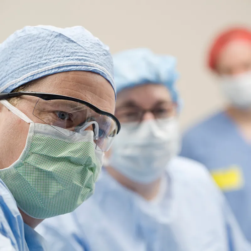 A Novant Health doctor is in surgery wearing personal protective equipment (PPE) including glasses and mask. Behind him are other team members, who are also dressed in PPE. 